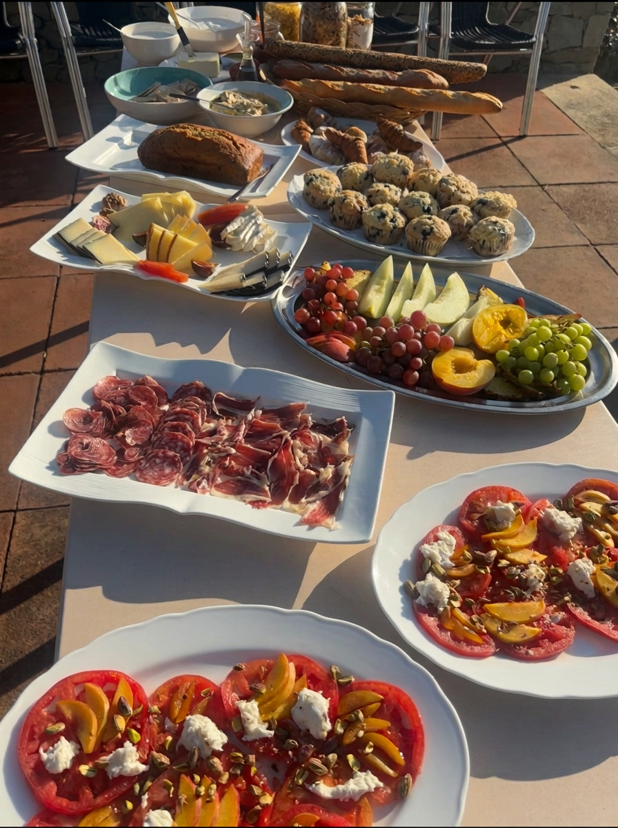 Full outdoor breakfast buffet table with breads, cheeses, charcuterie, fruit and tomato salads